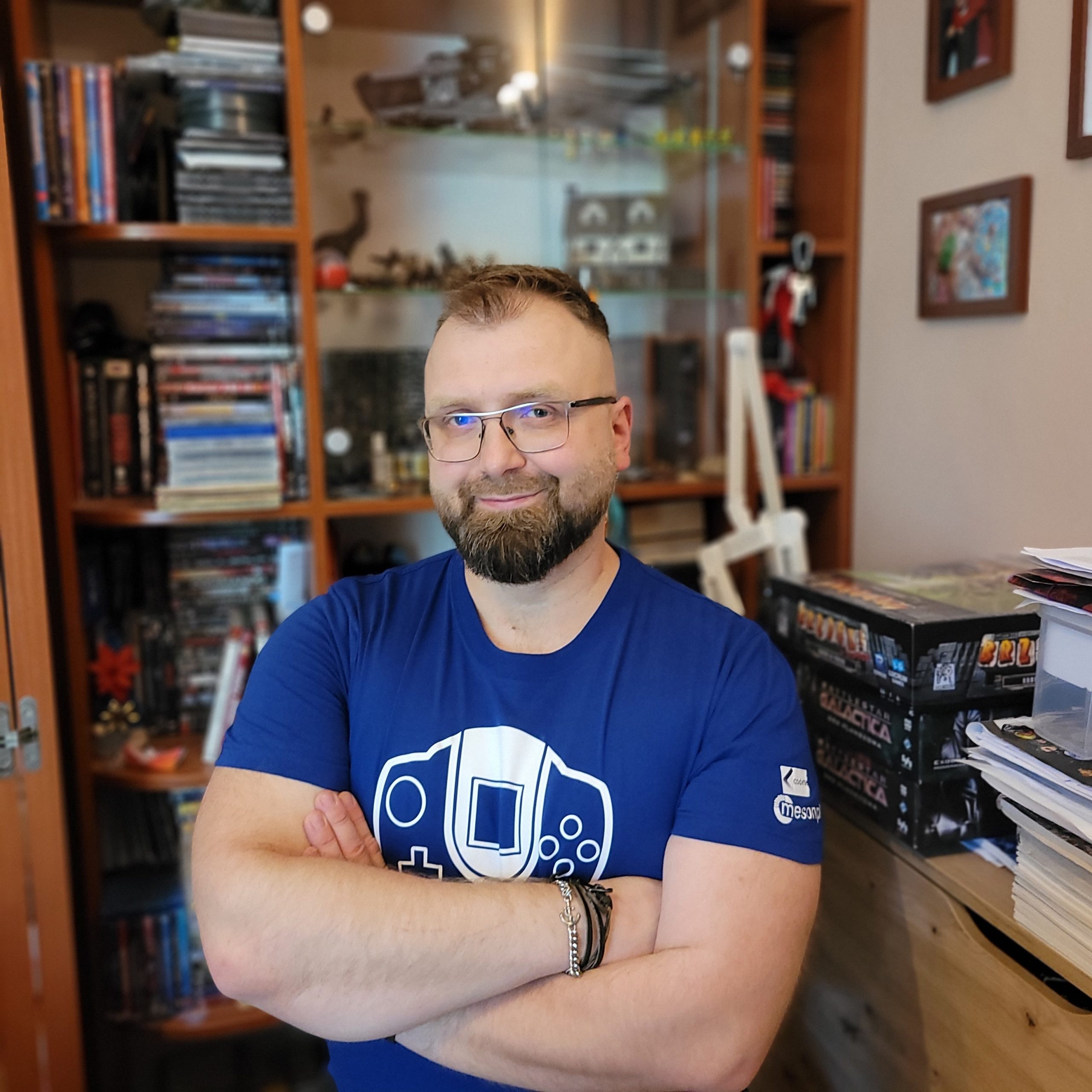 Zdjęcie przedstawia uśmiechniętego mężczyznę z brodą i okularami, stojącego ze skrzyżowanymi rękami. Ma na sobie ciemnoniebieską koszulkę z białym nadrukiem stylizowanego kontrolera do gier i napisem RetroSfera. W tle widać drewniany regał z książkami, płytami i innymi przedmiotami. Po prawej stronie zdjęcia widać fragment szafki i kartonowe pudełka.